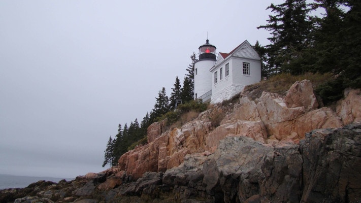 A small white lighthouse rises from a rocky shore and foggy ocean in Acadia National Park
