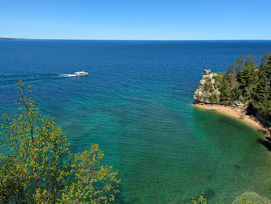 A white boat cruises by in the turquoise waters of Lake Superior. Golden sand and black and white cliffs make up the shoreline