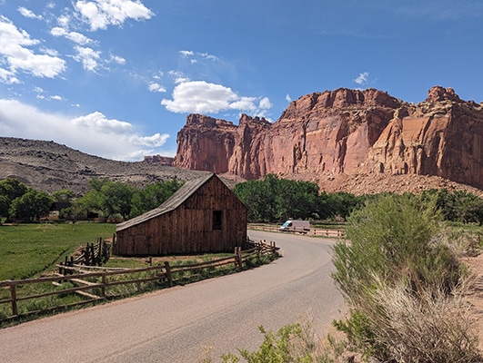 A wooden barn in green, irrigated fields rests below a cliff face of red rock