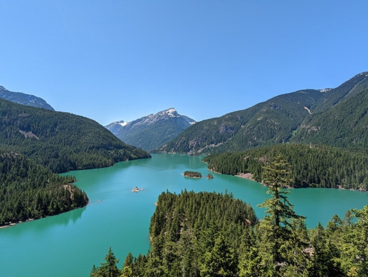 The vibrant blue-green water of Diablo Lake is surrounded by evergreen-covered mountains, with a snow-covered peak in the distance.