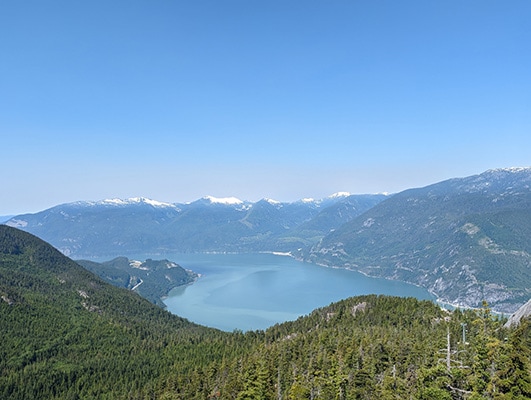 The blue water of the Howe Sound rests beyond tree-covered hills and below distant snow-covered peaks