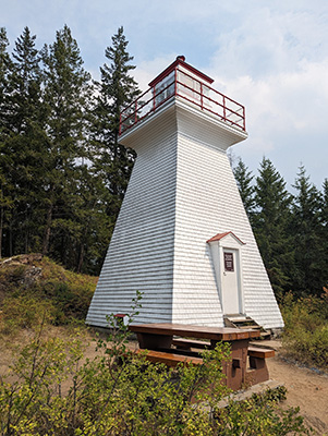 An almost pyramidal white lighthouse with red trim sits in front of evergreen trees