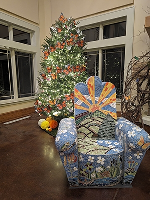 A Christmas tree decorated with monarch butterflies and a mosaic chair with a sun, path, and plants and animals