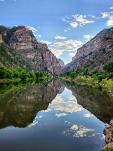 Towering rocky cliffs rise above a still river reflecting their images back to the blue, cloudy sky