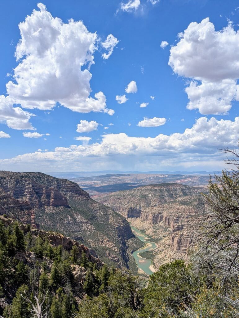 The Green River (which is actually green) flows through red, brush-covered hills beneath a blue sky and perfect white clouds