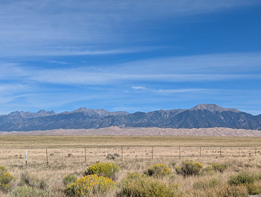 High sand dunes rise below towering rocky mountains and a blue skky