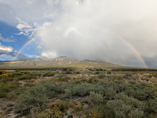 A bright rainbow rises above scrubland and rock-strewn mountains