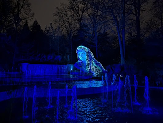 A female figure rises from the ground. She's covered in blue and white lights and situated behind a playful water fountain