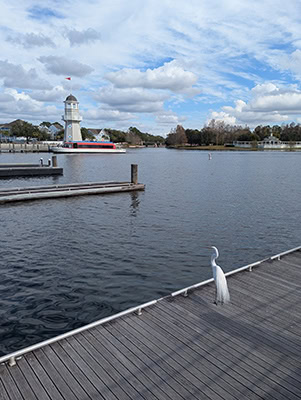 A great white egret on a dock appears to be looking at a small white lighthouse across a small lake