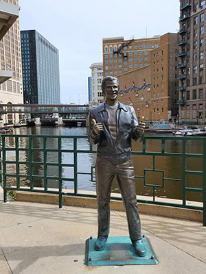 A bronze statue of a man giving two thumbs up. The man appears to be wearing a leather jacket, jeans, and has a slicked-back 1950s hairstyle