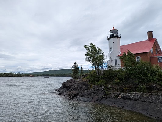 A white 2-story lighthouse and an attached keeper's cottage with a red roof preside over the gray waters and rocky shores of a little bay under cloudy skies