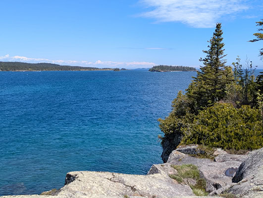 Gray rocks and green evergreen trees in the foreground give way to vibrant blue lake water, bluebird skies, and dark islands in the distance