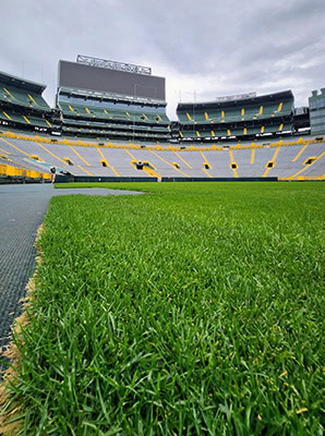 A picture taken from almost ground level of the green grass of a football stadium with the bleachers and a Jumbotron above