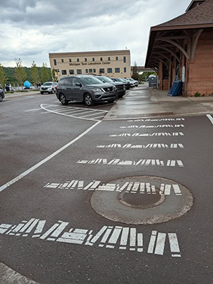 A white pedestrian crosswalk on a gray street with cars and buildings in the background. Instead of the white stripes found on most US crosswalks, this one has white rows of books