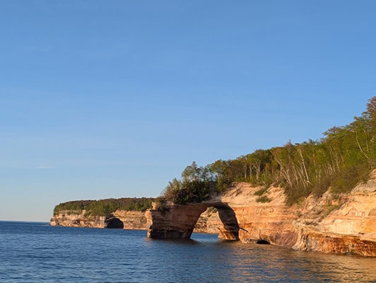 An arch in the yellow cliffs. The dark blue waters of a lake are below. The cliffs are mostly yellowish in the evening light but there are streaks of other colors present