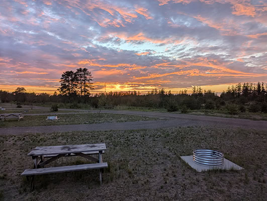 The golden sun is setting behind some silhouetted trees. The sky is glorious with clouds and sky in shade of purple, blue, and pink. A picnic table and fire pit are easily overlooked in the foreground
