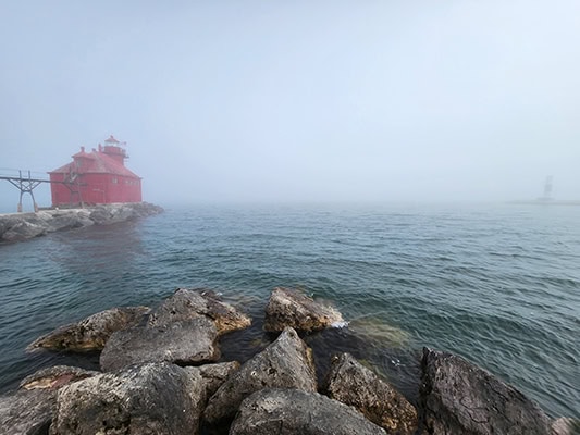 A red lighthouse is slightly obscured by fog on the left. Another structure, possibly a light beacon, has almost disappeared into the fog on the right. The center of the photo is moody blue water and dense fog, with wet stones in the foreground