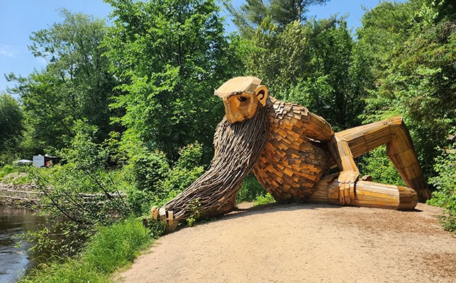A giant, unpainted wooden troll figure reclines on his right elbow. He's dangling the end of his beard in the river in an apparent effort to catch fish
