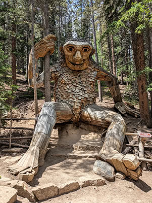 A giant troll, whose torso is at least twice as high as an average-sized adult, sits in the forest with his legs stretch before him. He holds on to the trunk of a tree in his right hand