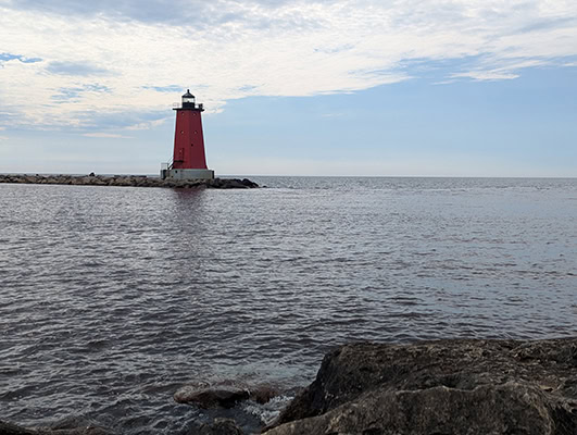 A short red lighthouse is at the end of a stone breakwater. A lake surrounds it. There are more rocks in the foreground. The sky is pale blue with some white cloud layers