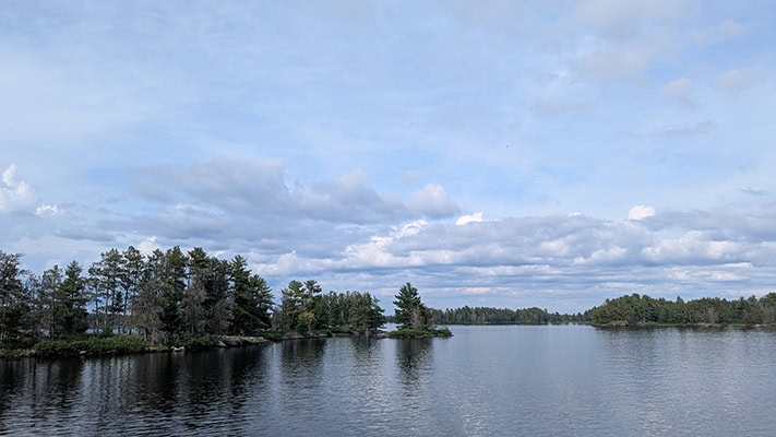 A blue lake stretches into the distance, dotted with islands and perhaps shoreline covered in evergreen trees. The sky is a soft blue with lots of white, blue, and gray clouds