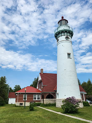 A white lighthouse rises above a low, red-roofed keeper's cottage. Trees and grass surround the structures, with no water in this view
