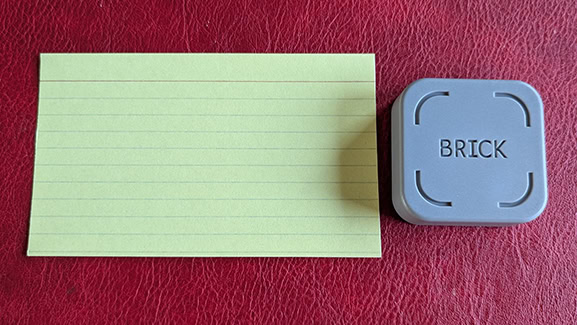 A square Brick device next to a yellow standard index card. The Brick is less than half the size of the card