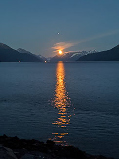 A yellow moon rises between some snow-covered peaks. The yellow light from the moon is reflected in the dark waters below